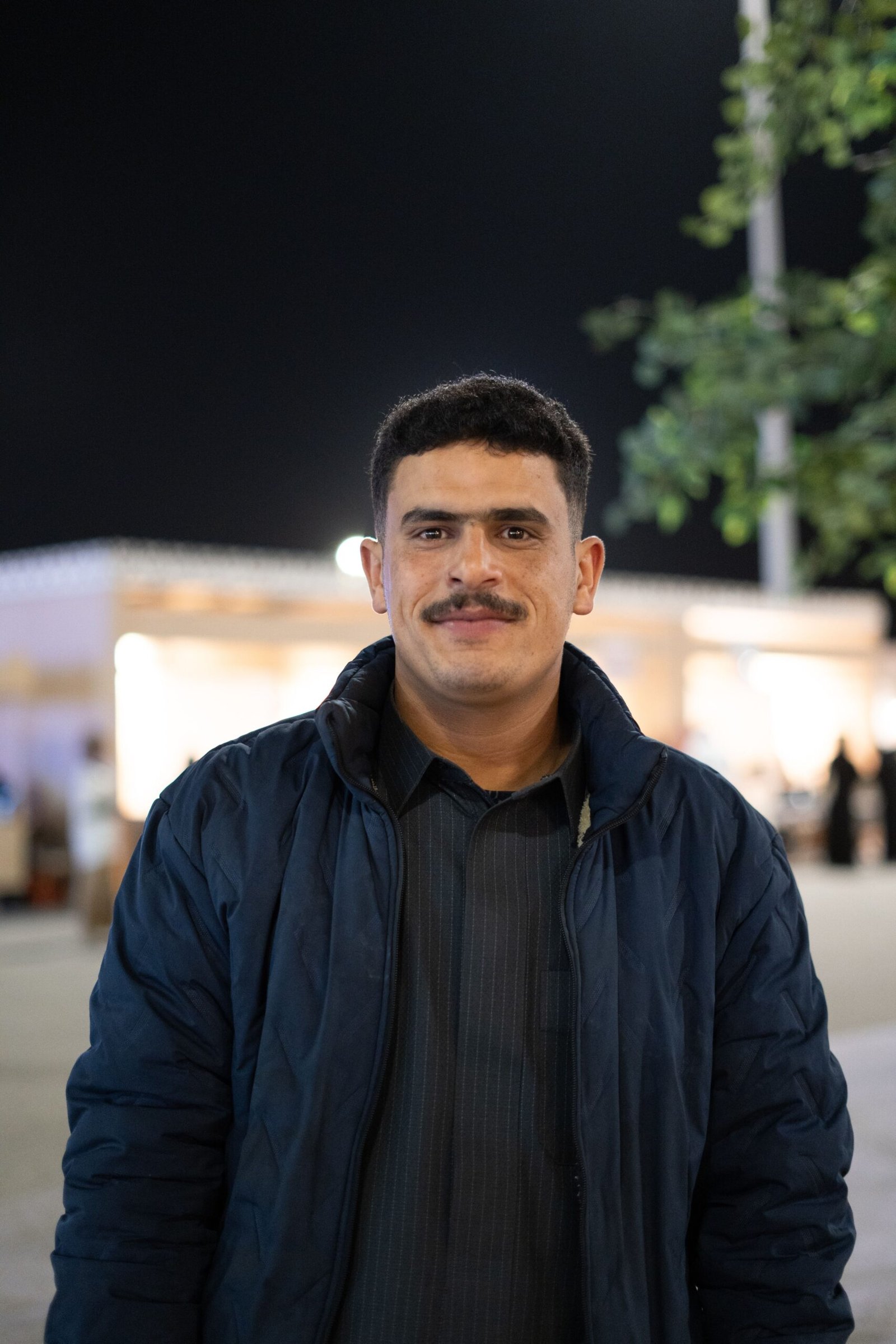 A cheerful man in a blue jacket smiles confidently during a night scene outdoors.
